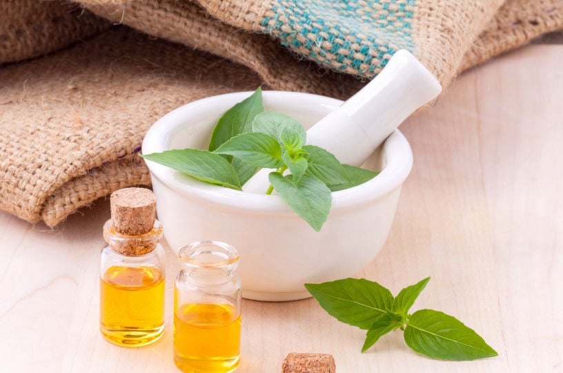 Mortar and pestle with fresh green herbs and small bottles of essential oil on a wooden surface